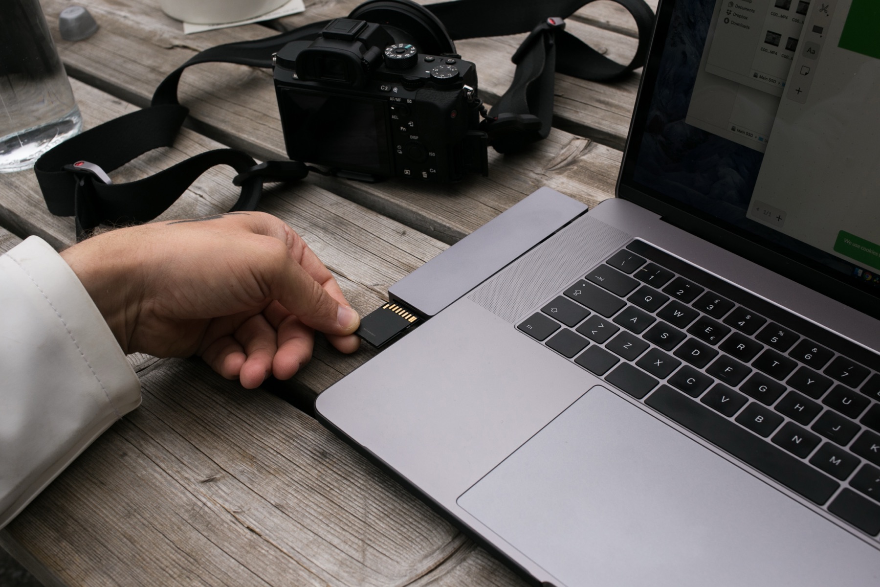 A hand slides an SD card into a laptop on a wooden desk, a camera beside it